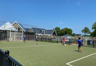 Kinderen spelen voetbal op een buitenveld bij een lodge, met huisjes en een speeltuin zichtbaar achteraan.