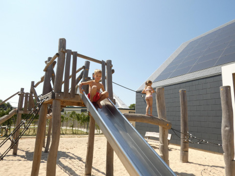 Kinder spielen auf einem Spielplatz mit Rutsche im Sand neben einer Lodge unter blauem Himmel.