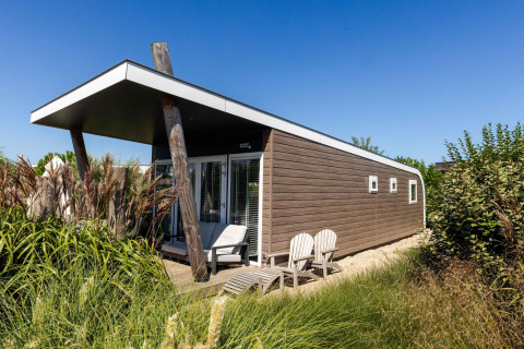 Moderno loft playero con wellness en Camping Zonneweelde, rodeado de vegetación bajo un cielo azul claro.
