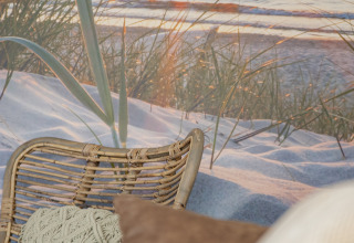 Afslappende loungeområde med fletstol, puder og strandbaggrund ved solnedgang, Camping Zonneweelde.