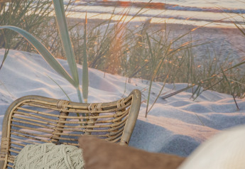 Afslappende loungeområde med fletstol, puder og strandbaggrund ved solnedgang, Camping Zonneweelde.