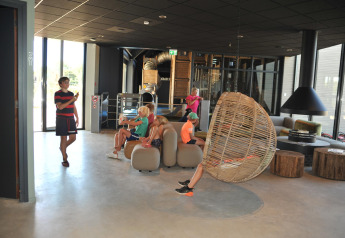 Børn slapper af i moderne lodge-lounge med store vinduer, hængestol og hyggelig indretning.