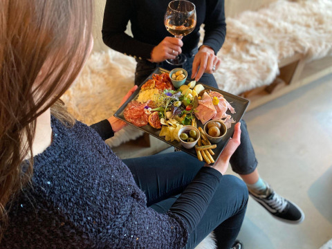 To personer sidder på en lodge med et glas vin og deler en charcuteriplade med snacks og pålæg.