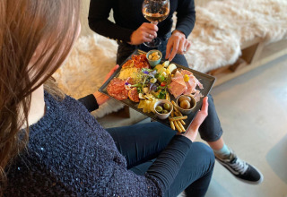 To personer sidder på en lodge med et glas vin og deler en charcuteriplade med snacks og pålæg.