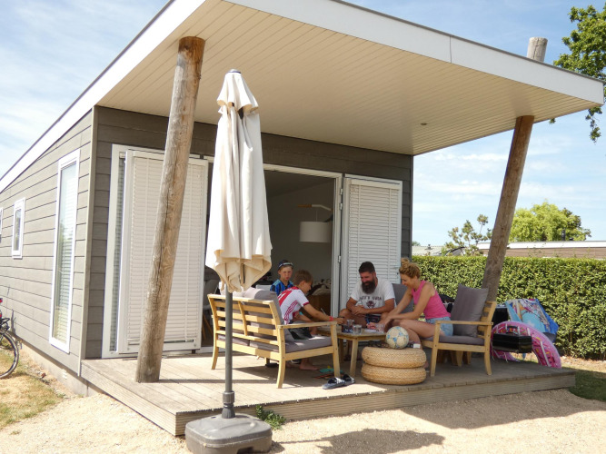 Family relaxes on the porch of a Beach Loft lodge at Camping Zonneweelde in the Netherlands, summertime scene.
