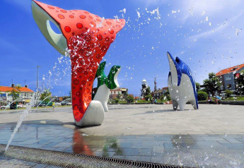 Esculturas coloridas de animales marinos y chorros de fuente en una plaza con edificios y cielo azul.