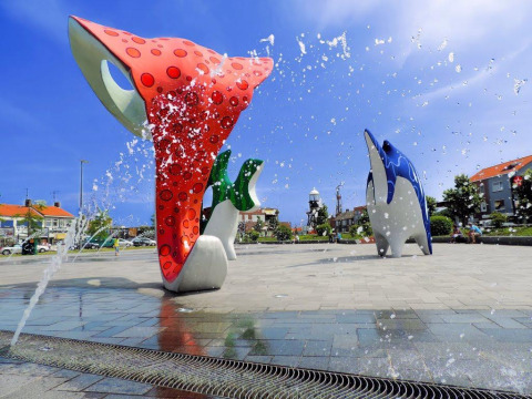 Sculptures colorées d’animaux marins et jets de fontaine sur une place, entourée d’immeubles et ciel bleu.