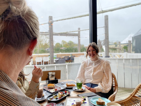Dos mujeres disfrutan pasteles y bebidas en una acogedora cabaña con ventanas grandes hacia la terraza.