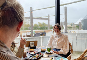 Two women enjoy cakes and drinks in a cozy lodge with large windows overlooking the outdoor terrace.