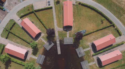 Vista aérea de alojamiento glamping con seis cabañas techadas, senderos, setos y muelles junto a un lago.