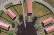 Luchtfoto van een glamping met zes huisjes met rood dak, paden, heggen en vier steigers aan het water.