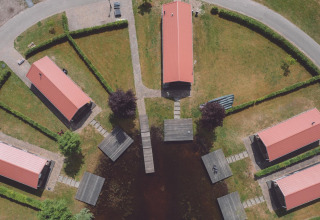 Vista aérea de alojamiento glamping con seis cabañas techadas, senderos, setos y muelles junto a un lago.