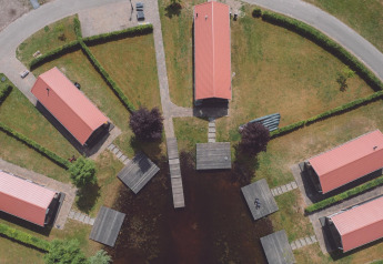 Luftfoto af glamping-område med seks rødtags hytter, stier, hække og små bådebroer ved en sø.