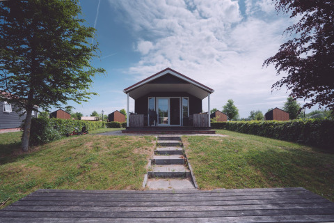 Foto di un alloggio glamping moderno con veranda in legno, prato, alberi e cielo parzialmente nuvoloso.