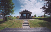 Foto di un alloggio glamping moderno con veranda in legno, prato, alberi e cielo parzialmente nuvoloso.