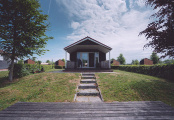 Foto van een glampingaccommodatie met houten terras, groene tuin en bomen onder een wisselvallige lucht.