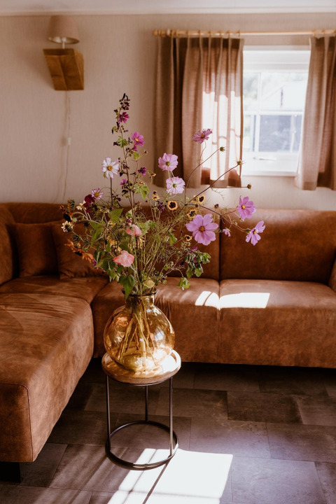 Blomsterbuket i glasvase på bord i brun stue med hjørnesofa, sollys og vindue i Hollandsk feriepark.