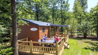 Familie sidder udenfor på terrassen ved Cottage på Veluwecamping De Pampel, omgivet af frodig skov.