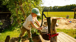Niño usando una bomba de agua antigua, bajo el sol, cerca del parque infantil de Feather Down De Lange Weide.