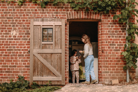Two people stand in a barn doorway of a brick building with a wooden door and climbing plants.