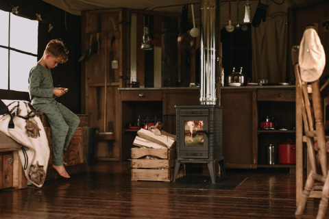 Boy in pajamas sits on a bed in a cozy glamping cabin with a wood stove and rustic decor.