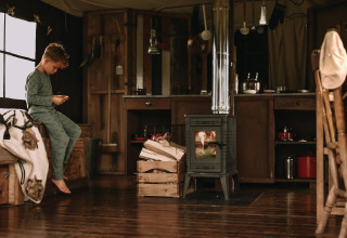 Dreng i pyjamas sidder på seng i hyggelig glamping-hytte med brændeovn og landlig indretning.