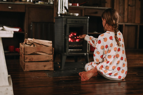 Et barn i pyjamas sidder foran en brændeovn i en hytte, omgivet af træstykker og hyggelig atmosfære.