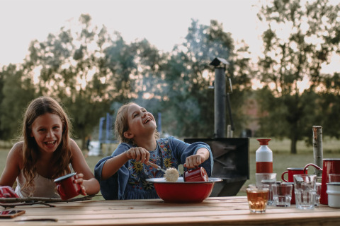 Zwei lachende Kinder essen im Freien an einem Holztisch im Ferienpark Landgoed ter Wijnendale, Belgien.