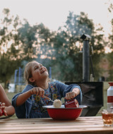 To glade børn spiser sammen udendørs ved et træbord i aftensol i Landgoed ter Wijnendale, Belgien.