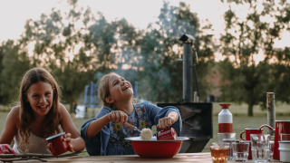 Deux enfants souriants partagent un repas en plein air à une table en bois à Landgoed ter Wijnendale, Belgique.