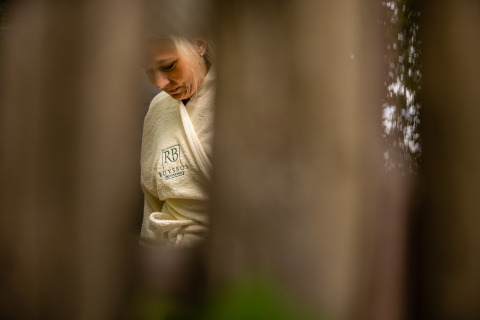 Mujer en bata con logo Ruysbos, vista a través de troncos borrosos en Boshuys, Buitengoed Ruysbos, Países Bajos.