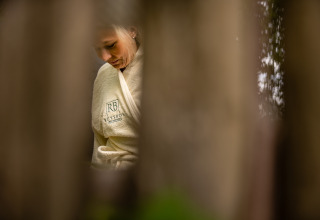 Mujer en bata con logo Ruysbos, vista a través de troncos borrosos en Boshuys, Buitengoed Ruysbos, Países Bajos.