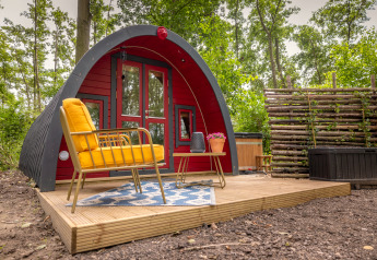 Bogengeformtes Tiny House mit roter Fassade, gelbem Sessel und Terrasse im Buitengoed Ruysbos, Niederlande.