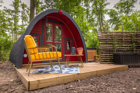 Træhytte med buet tag, rød facade, gul stol og terrasse omgivet af træer i Buitengoed Ruysbos, Holland.
