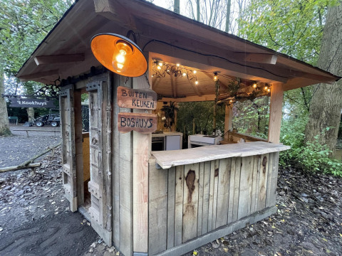 Una pequeña casa de madera con cocina al aire libre, carteles de madera y luces, en un área boscosa.