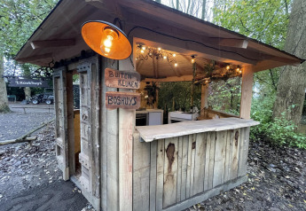 Una pequeña casa de madera con cocina al aire libre, carteles de madera y luces, en un área boscosa.
