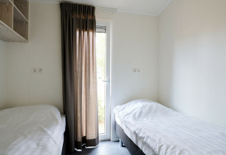 To enkeltsenge i et lyst værelse med en dør og brune gardiner i Sand Lodge, Holiday Park De IJsvogel.