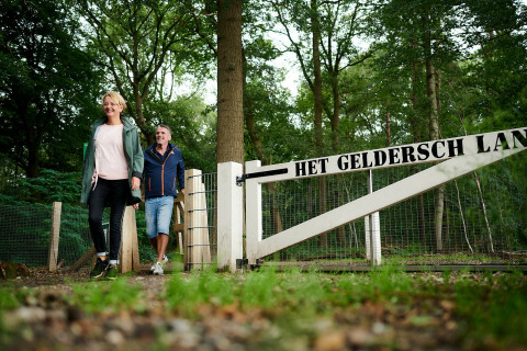 Dos personas pasan por una puerta de bosque con el letrero 'Het Geldersch Land' en Holiday Park De IJsvogel.