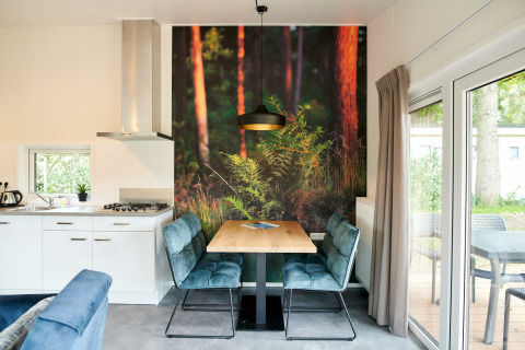 Tiny house moderne avec coin repas, fresque murale forêt, cuisine ouverte et grandes baies vitrées sur terrasse.