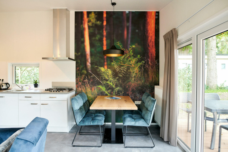 Modernes Tiny House mit kleiner Essecke, Waldmotiv an der Wand, offener Küche und großen Fenstern zur Terrasse.