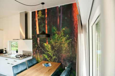 Moderne tiny house-keuken met houten tafel, blauwe stoelen en een bosmuurdecor naast grote ramen.