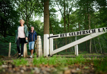 Dos personas cruzan una puerta con el letrero 'Het Geldersch Land' junto a una tiny house en el bosque.