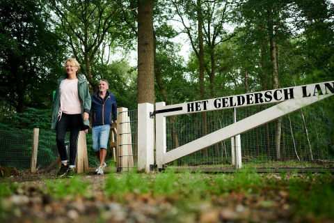 Twee mensen lopen door een hek met 'Het Geldersch Land' bij een tiny house in een bosrijke omgeving.