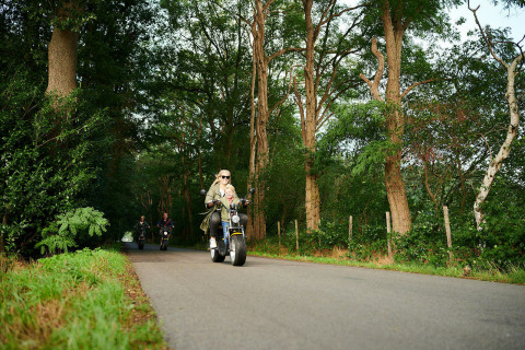 Mensen rijden met scooters over een landweg, omgeven door bomen, dicht bij een tiny house.