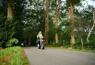 Personas paseando en scooter por un camino arbolado cerca de una tiny house, rodeados de naturaleza.