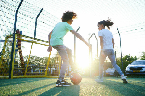 Dos niños juegan al fútbol en un campo exterior al atardecer, con coches y vallas al fondo y sol brillante.
