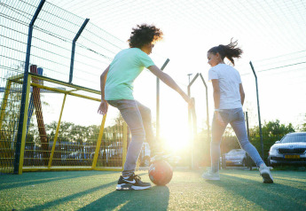 Two kids are playing soccer on an outdoor field at sunset, with parked cars and wire fencing in the background.