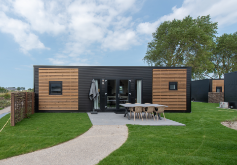 Cabaña moderna con fachada negra y madera, área de comedor exterior y césped verde bien cuidado al frente.