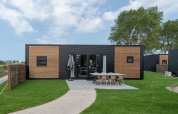 Cabaña moderna con fachada negra y madera, área de comedor exterior y césped verde bien cuidado al frente.