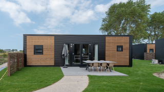 Cabaña moderna con fachada negra y madera, área de comedor exterior y césped verde bien cuidado al frente.